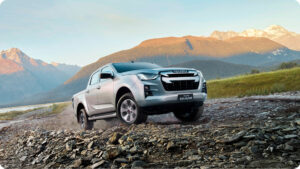 Silver Isuzu D-Max pickup driving over a rocky off-road terrain with a mountain range in the background.